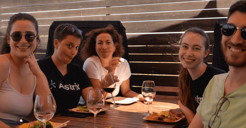 Astrix transforms non-human identity management, securing service accounts and enhancing NHI security. Alt text: Group of five enjoying a meal outdoors at a wooden table with wine glasses, seated against a wooden slat backdrop.