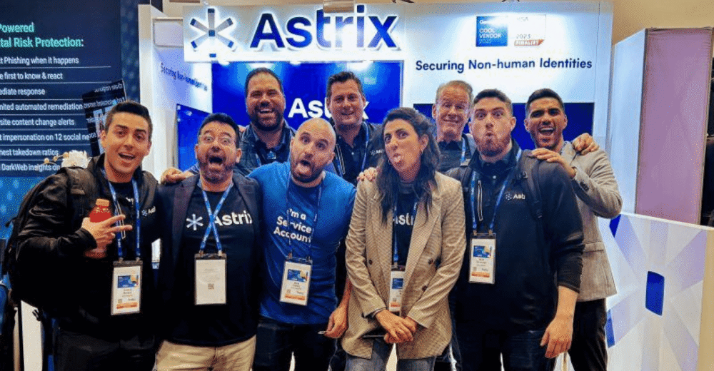 A diverse group of nine individuals in casual and business attire pose playfully in front of an Astrix booth at a conference, all wearing conference badges. Astrix is a leading NHI security platform.